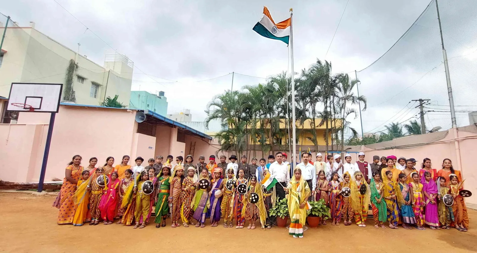 Large group of students and staff standing outdoors around the flag mast for an Independence Day celebration
