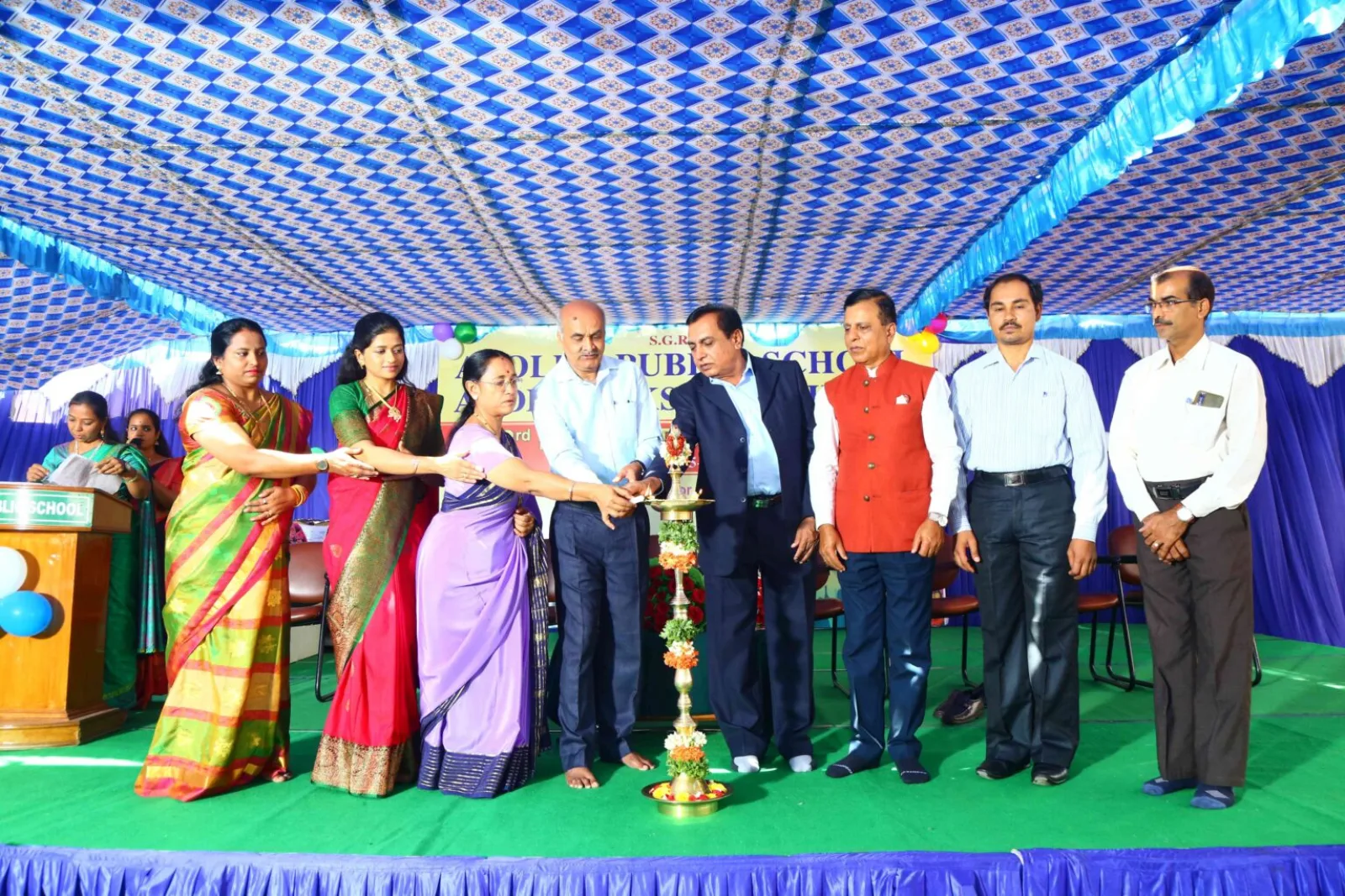 School management and guests lighting a ceremonial lamp on stage under a decorated blue tent