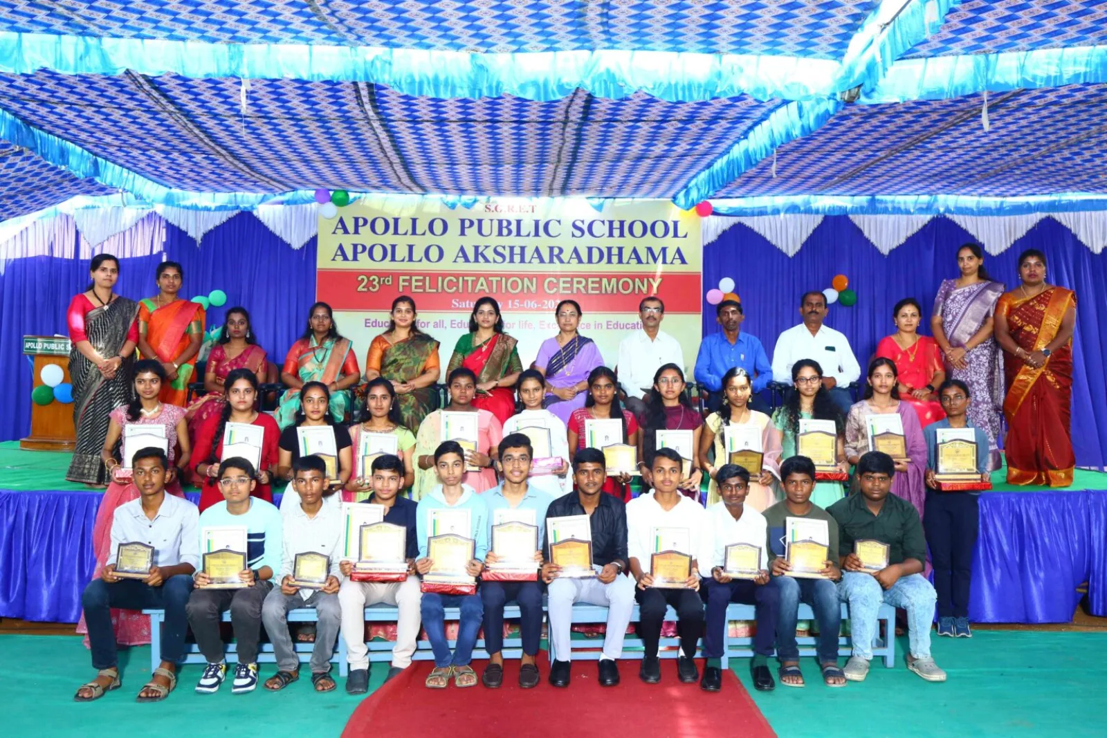 A group of students seated together holding certificates at the Apollo Aksharadhama branch, with a school banner behind them